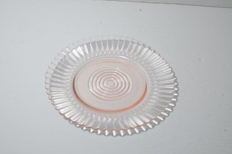 Puede incluir: Un plato de vidrio transparente con un centro rosa y un borde decorativo y acanalado. El centro del plato presenta c&iacute;rculos conc&eacute;ntricos. El plato es probablemente para servir alimentos.