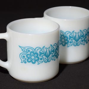 May include: Two white ceramic mugs with a turquoise floral design. One mug has a handle, and the other is handle-less. The floral pattern wraps around the mugs, creating a vintage aesthetic. The mugs are set against a dark background.