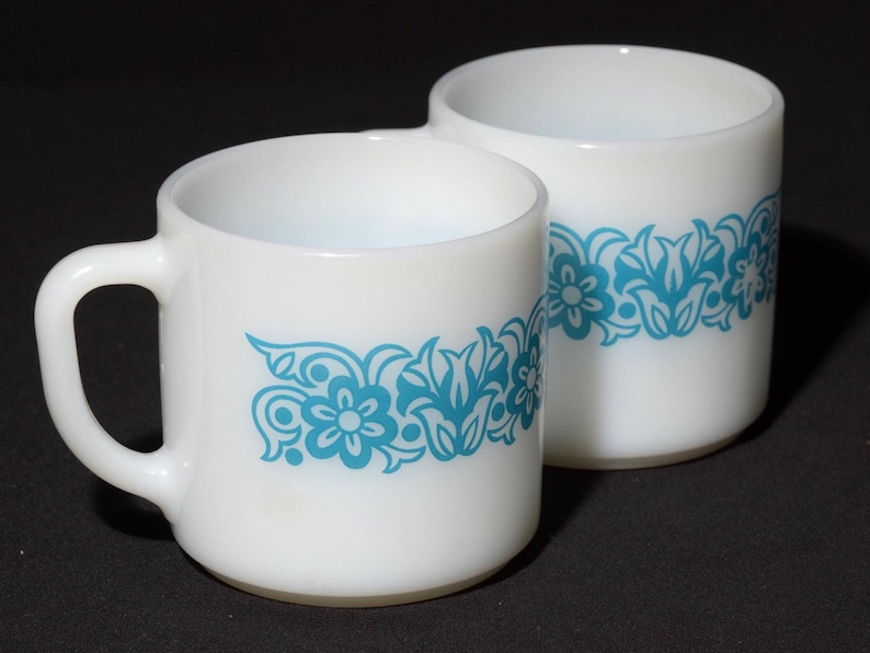 May include: Two white ceramic mugs with a turquoise floral design. One mug has a handle, and the other is handle-less. The floral pattern wraps around the mugs, creating a vintage aesthetic. The mugs are set against a dark background.