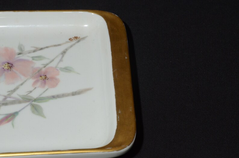 May include: Rectangular white ceramic tray with a gold-painted border. The tray features a floral design with pink flowers, green leaves, and brown stems. The corner of the tray has the initials "C.R." painted on it.