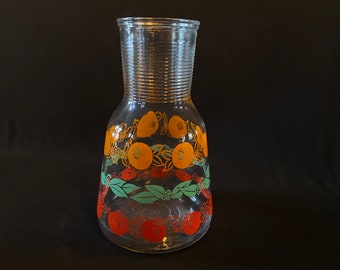 May include: A clear glass pitcher with a textured design. The pitcher has a wide base and a narrow neck. It features a colorful pattern of orange, red, and green fruit and leaves.