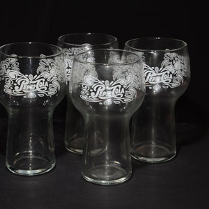 May include: Four clear glass tumblers with a white etched design of the Pepsi-Cola logo. The glasses have a wide base and a rounded top.