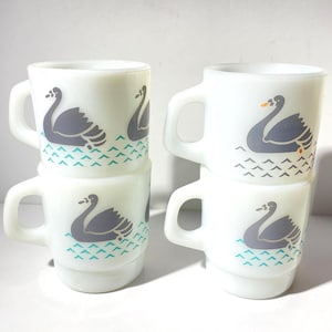 Könnte beinhalten: Vier weiße Keramikbecher gestapelt, jeder mit einem grauen Schwanenmuster auf einem türkis-weißen Wassermuster. Die Becher haben einen Henkel und einen abgerundeten Boden. Das Schwanenmuster ist ein herausragendes Merkmal.