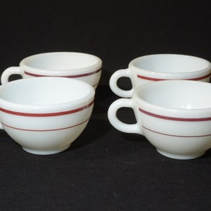 May include: Four white ceramic coffee mugs with a single red stripe around the top. The mugs are arranged in a square formation.