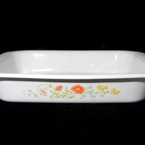 Peut inclure: Un plat de cuisson rectangulaire blanc aux coins arrondis et au bord large. L'extérieur présente un motif floral avec des fleurs orange, jaunes et bleues ainsi que des feuilles vertes centrées sur la longueur de la surface blanche.