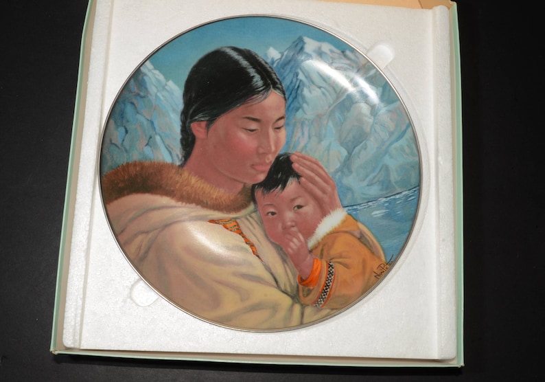 May include: A decorative plate depicting a mother and child in traditional attire, with a backdrop of snow-covered mountains and water. The plate has a silver rim and is presented in a protective box.
