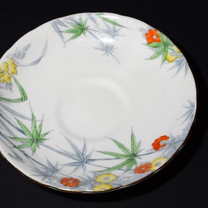 May include: White ceramic saucer with a floral design. The saucer features a gold rim and is decorated with green, yellow, and orange flowers and leaves. The design is set against a backdrop of gray bamboo-like leaves.