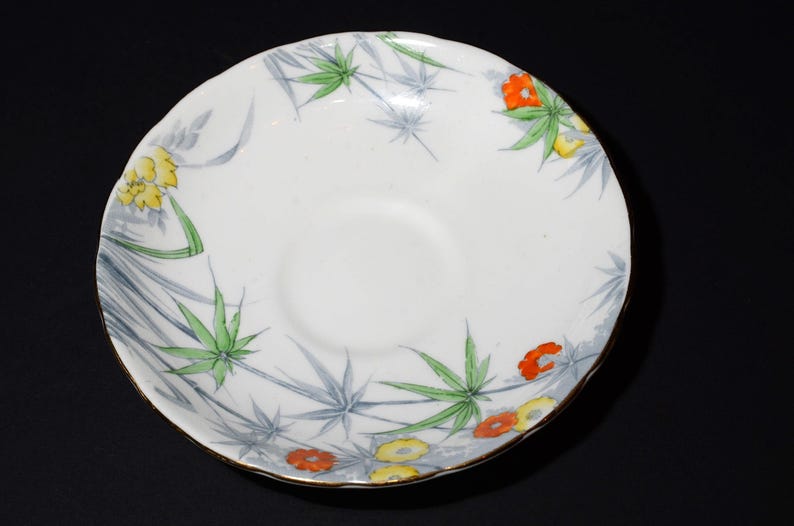 May include: White ceramic saucer with a floral design. The saucer features a gold rim and is decorated with green, yellow, and orange flowers and leaves. The design is set against a backdrop of gray bamboo-like leaves.