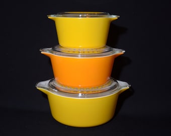 May include: A set of three vintage glass casserole dishes with lids. The dishes are stacked, with the top dish being yellow, the middle dish orange, and the bottom dish yellow.