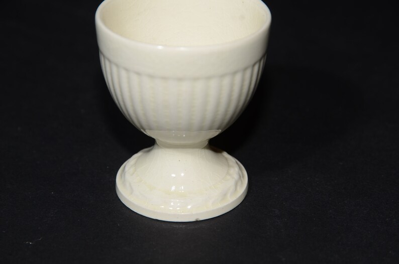 May include: A white ceramic egg cup with a fluted design and a pedestal base.