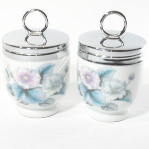 May include: Two white ceramic egg coddlers with floral designs and silver lids. Each lid has a ring handle.
