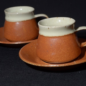 May include: Two brown and white ceramic coffee cups with matching saucers. The cups have a white band around the top and a brown glaze on the bottom.
