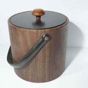 Op de afbeelding: Een houten ijs emmer met een zwart leren handvat en een houten deksel met een knop. De emmer is bruin en heeft een natuurlijke houtnerf.