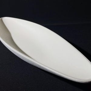 May include: A white ceramic serving dish shaped like a long, narrow boat. The dish has a smooth, slightly textured surface.