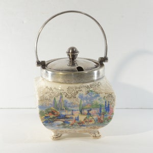 Op de afbeelding: Een vintage keramische suikerpot met een zilverkleurig deksel en handvat. De pot heeft een handgeschilderde landschapsscène met bomen, water en figuren. De pot staat op drie kleine voetjes.