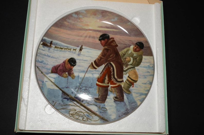 May include: A ceramic plate with a colorful painting of three people in winter clothing building an igloo. The scene is set against a snowy landscape with a sunset in the background.