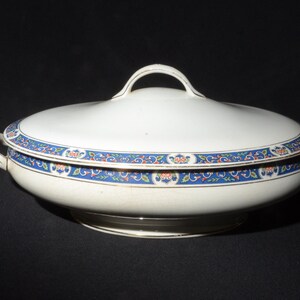 May include: A white ceramic serving dish with a lid and handles. The dish has a blue and red floral pattern around the rim. The lid has a loop handle. The dish is on a black background.