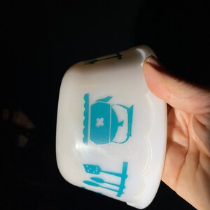 May include: White ceramic bowl with a scalloped edge and a blue design of kitchen utensils and a teapot. The teapot has a cross on the lid.