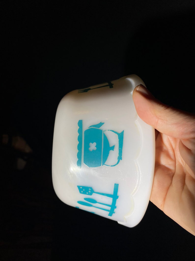 May include: White ceramic bowl with a scalloped edge and a blue design of kitchen utensils and a teapot. The teapot has a cross on the lid.