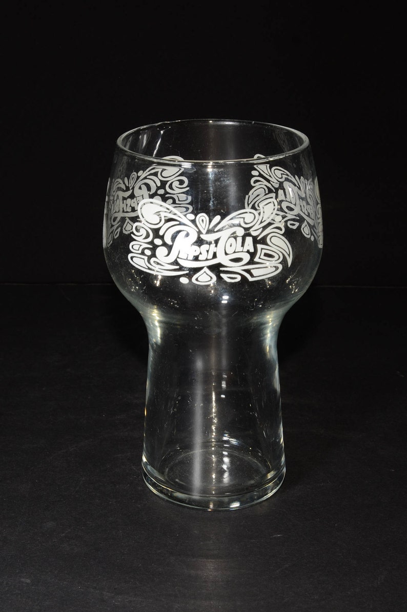 May include: A clear glass drinking glass with a wide base and a flared top. The glass has a white etched design of the Pepsi-Cola logo and swirls.