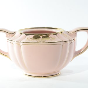 May include: A pink sugar bowl with gold trim and a floral design. The bowl has two handles and a rounded shape.