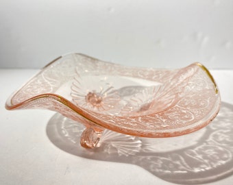 May include: A pink glass bowl with a scalloped edge and a floral design. The bowl has a gold rim and three feet.