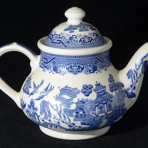Puede incluir: Una tetera de cerámica blanca y azul con tapa. La tetera presenta un patrón Willow clásico con ilustraciones azules detalladas de pagodas, pájaros y paisajes. El asa y el pico son curvos, y la tapa tiene un pequeño pomo.
