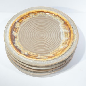 Peut inclure: Une pile de six assiettes en céramique avec un glaçage brun et blanc. Les assiettes ont un motif en spirale au centre et un bord brun.