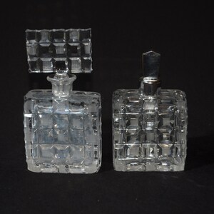 May include: Two clear glass perfume bottles with square, textured designs. The bottle on the left has a square stopper, while the bottle on the right has a silver stopper.