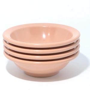 May include: Four light pink ceramic bowls are stacked. Each bowl has a rounded base and a wide, flat rim. The bowls are arranged one on top of the other, with the rims facing upwards. The background is white.
