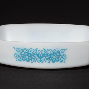 Könnte beinhalten: Eine weiße, rechteckige Backform aus Glas mit abgerundeten Ecken und glatter Oberfläche. In der Mitte der Vorderseite befindet sich ein dekoratives, cyanblaues Blumenmuster mit symmetrischen Ranken und zwei stilisierten Blüten.
