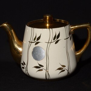 May include: A white teapot with a gold lid and handle. The teapot features a gold leaf design and a silver sticker on the side.