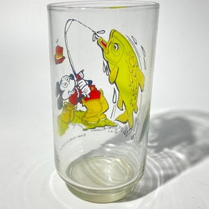 K&ouml;nnte beinhalten: Ein klares Glas mit einer Cartoon-Illustration von Mickey Mouse beim Angeln. Mickey, in roten Shorts und einem gelben Hemd, zieht einen gro&szlig;en gelben Fisch an Land. Das Glas hat einen wei&szlig;en Boden und einen leicht ausgestellten Rand.