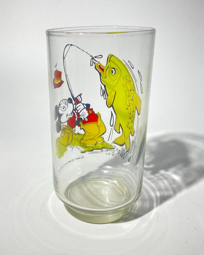 K&ouml;nnte beinhalten: Ein klares Glas mit einer Cartoon-Illustration von Mickey Mouse beim Angeln. Mickey, in roten Shorts und einem gelben Hemd, zieht einen gro&szlig;en gelben Fisch an Land. Das Glas hat einen wei&szlig;en Boden und einen leicht ausgestellten Rand.