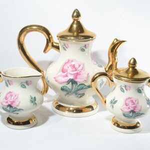 May include: A white porcelain tea set with pink roses and gold accents. The set includes a teapot, creamer, and sugar bowl.