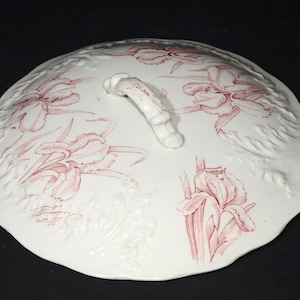 Puede incluir: Tapa de cerámica blanca con un diseño floral rosa y un asa. La tapa está estampada con un patrón floral y tiene un borde elevado.