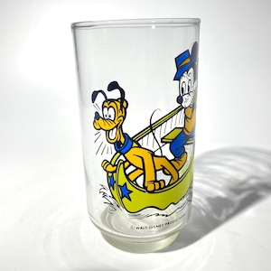 May include: Clear glass tumbler featuring a colorful cartoon illustration of Mickey Mouse and Pluto in a yellow boat. The characters are depicted in their classic outfits. The bottom of the glass reads "&copy; WALT DISNEY PRODUCTIONS."