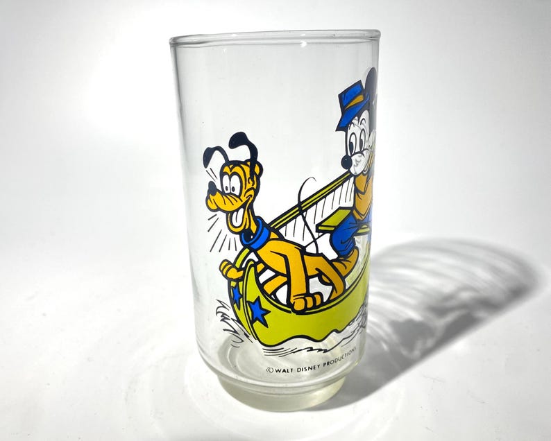 May include: Clear glass tumbler featuring a colorful cartoon illustration of Mickey Mouse and Pluto in a yellow boat. The characters are depicted in their classic outfits. The bottom of the glass reads "&copy; WALT DISNEY PRODUCTIONS."
