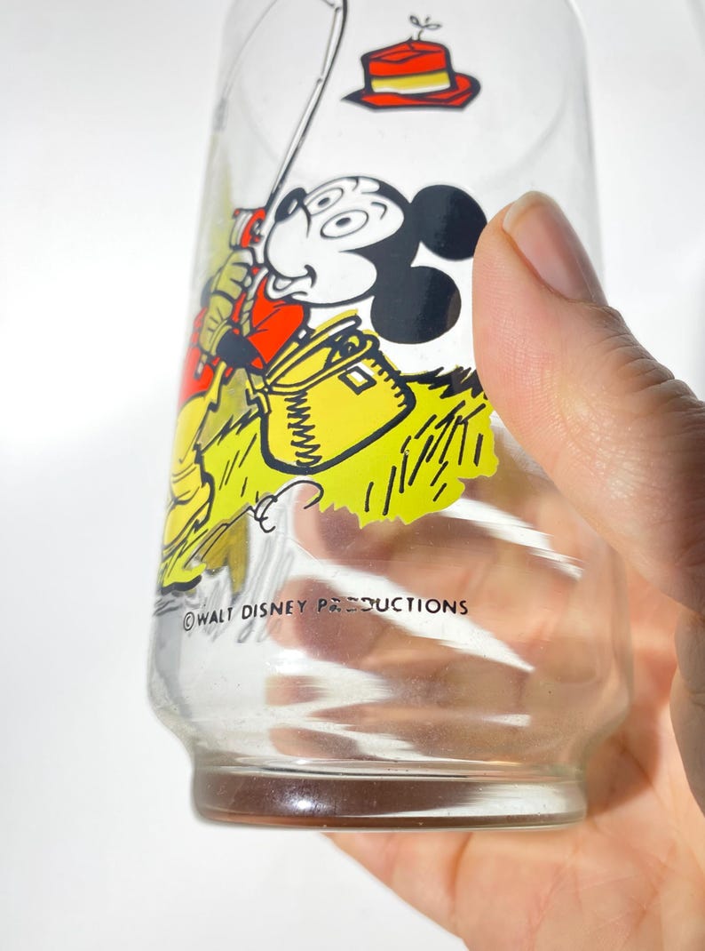 K&ouml;nnte beinhalten: Klares Glas mit einer Vintage-Mickey-Mouse-Angelszene. Mickey ist in einem roten Hemd und gelben Hosen dargestellt und h&auml;lt eine Angelrute. Das Glas hat einen gelb-gr&uuml;nen Hintergrund mit dem Text "WALT DISNEY PRODUCTIONS".