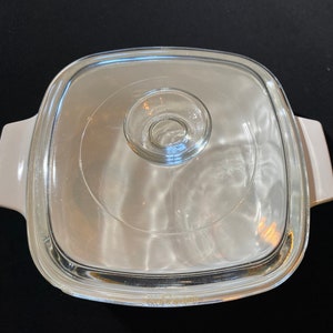 K&ouml;nnte beinhalten: Wei&szlig;e quadratische Auflaufform mit einem Glasdeckel. Der Deckel hat einen kleinen Knopf in der Mitte.