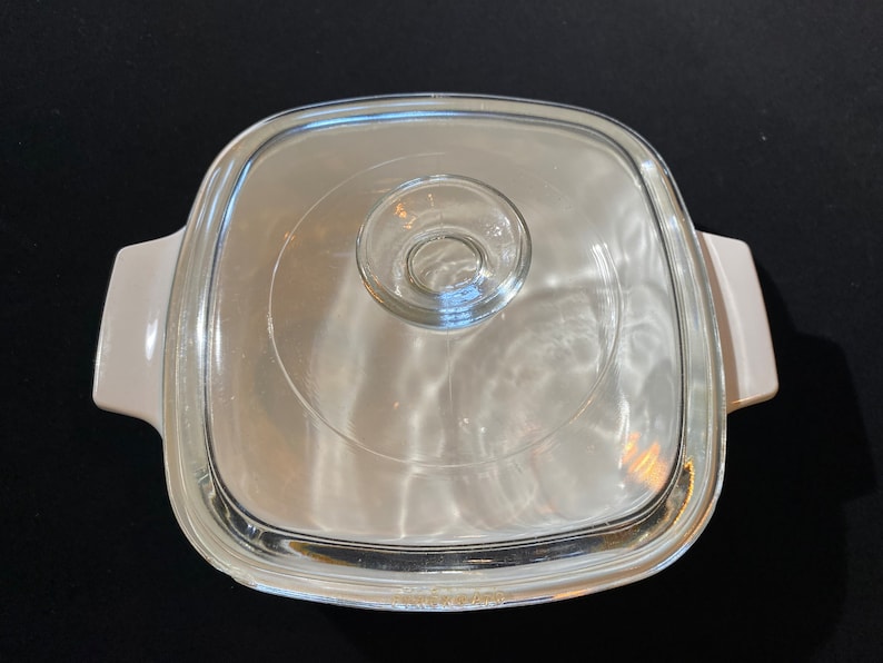 K&ouml;nnte beinhalten: Wei&szlig;e quadratische Auflaufform mit einem Glasdeckel. Der Deckel hat einen kleinen Knopf in der Mitte.