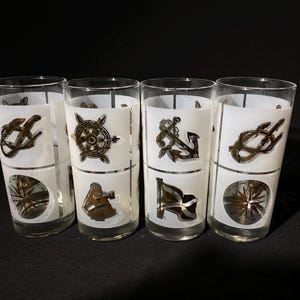 以下が含まれることがあります： 白と金の航海をテーマにしたデザインが施された透明なガラス製のタンブラー4個セット。各タンブラーには、コンパス、錨、船の車輪、結び目など、異なるデザインが施されています。