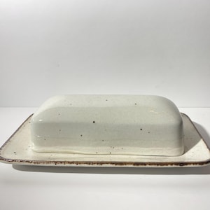 Peut inclure: Un beurrier en céramique blanche avec un glaçage moucheté et un bord brun. Le plat a une base rectangulaire et un dessus arrondi.