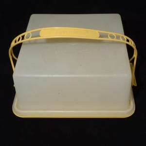 Peut inclure: Un porte-gâteau Tupperware vintage. Le récipient est rectangulaire avec une base et un couvercle jaune pâle. Il possède une poignée jaune assortie avec le logo Tupperware. Le récipient est en plastique translucide.
