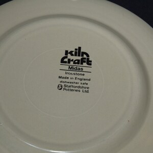 May include: Close-up of the bottom of a white ceramic plate with black text that reads "kiln Craft Midas Ironstone Made in England dishwasher safe Staffordshire Potteries Ltd."