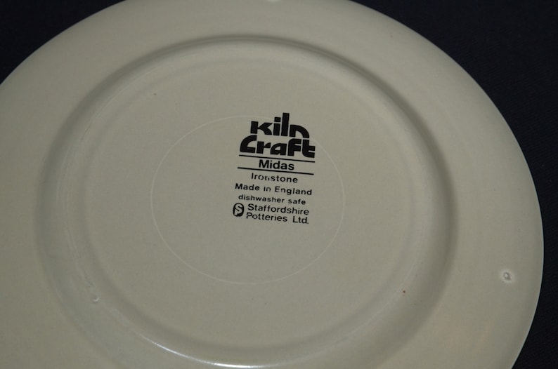 May include: Close-up of the bottom of a white ceramic plate with black text that reads "kiln Craft Midas Ironstone Made in England dishwasher safe Staffordshire Potteries Ltd."