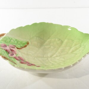 May include: A green ceramic bowl shaped like a leaf with pink flowers and green stems. The bowl is designed to look like a leaf with a textured surface.