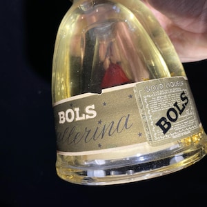 May include: A clear glass bottle of Bols Ballerina gold liqueur. The bottle has a gold label with the Bols logo and the words "Ballerina" and "Gold Liqueur". The bottle contains a golden liquid with gold flakes and a red object.