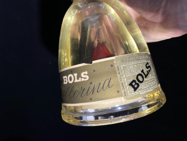May include: A clear glass bottle of Bols Ballerina gold liqueur. The bottle has a gold label with the Bols logo and the words "Ballerina" and "Gold Liqueur". The bottle contains a golden liquid with gold flakes and a red object.