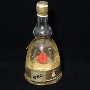 May include: A vintage Bols bottle with a clear glass body and a gold-colored cap. The bottle contains a red decorative element and gold flakes suspended in a yellow liquid. The label reads "Bols".